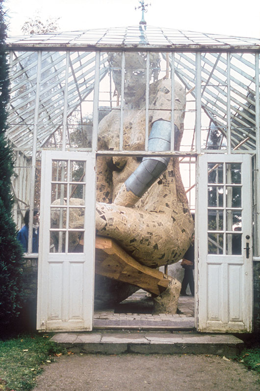 Artwork by Susanne Ussing. A giant statue of a woman squatting inside a greenhouse with walls and roof of glass. The statue is made of papier mâché, sheets of metal, and wood. The woman has one shoulder raised and looks downward, solemnly.